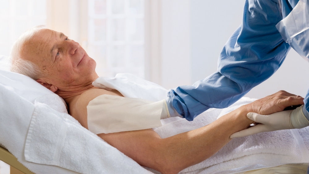 Männlicher Patient im Bett mit Krankenschwester, die an seinem Oberarm eine Körperdekolonisation durchführt