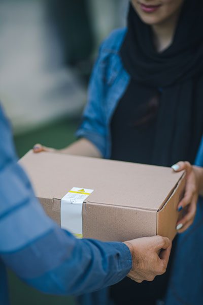 Eine Dame nimmt ein braunes, mittelgroßes Paket von einem Postboten entgegen. 