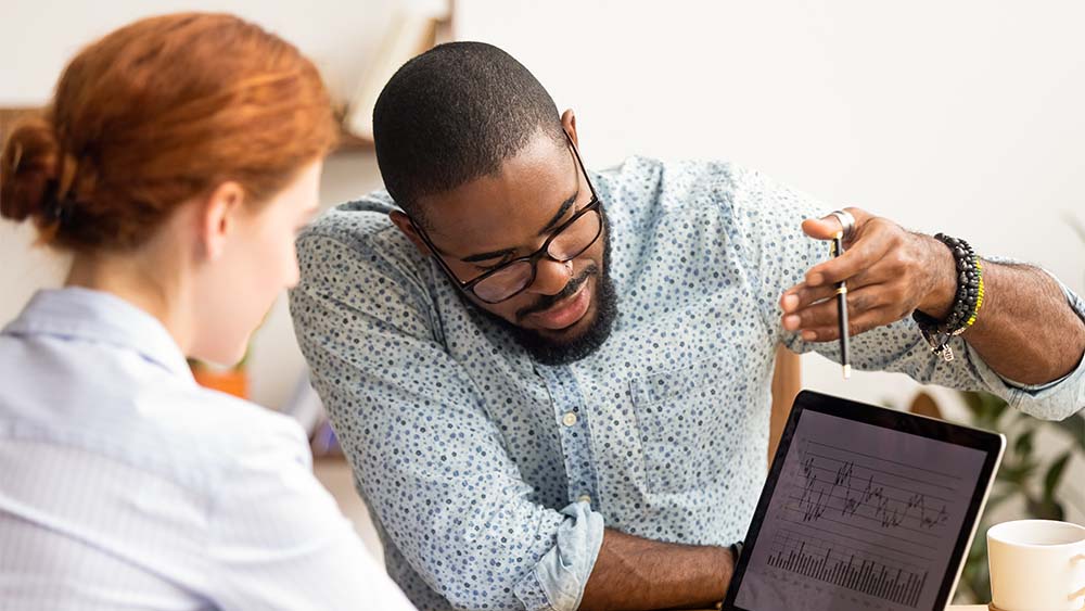 Zwei Personen diskutieren Geschäftsgrafiken und Diagramme auf einem Tablet in einem Büroraum. 