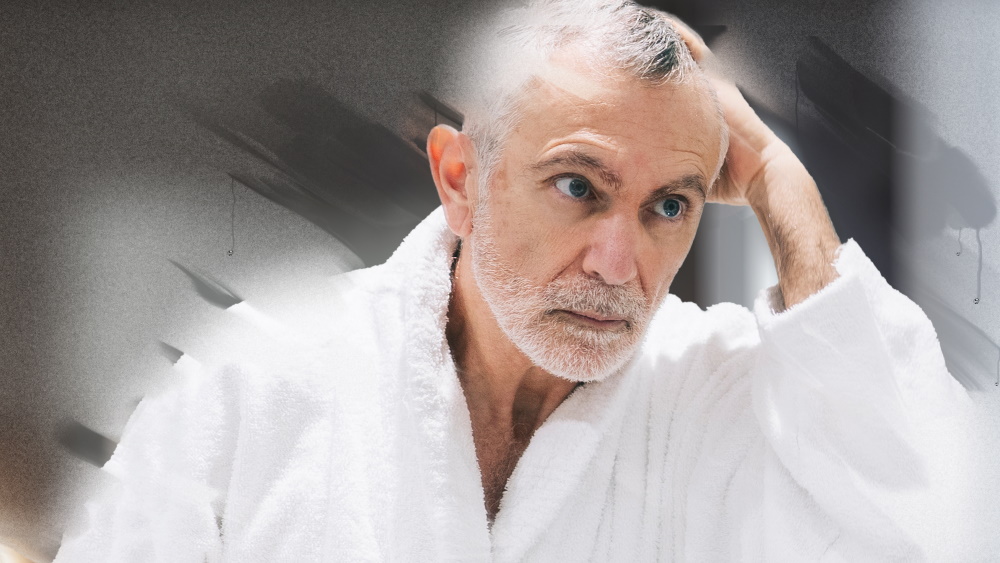 Älterer Mann im Bademantel schaut nachdenklich in beschlagenen Badezimmerspiegel