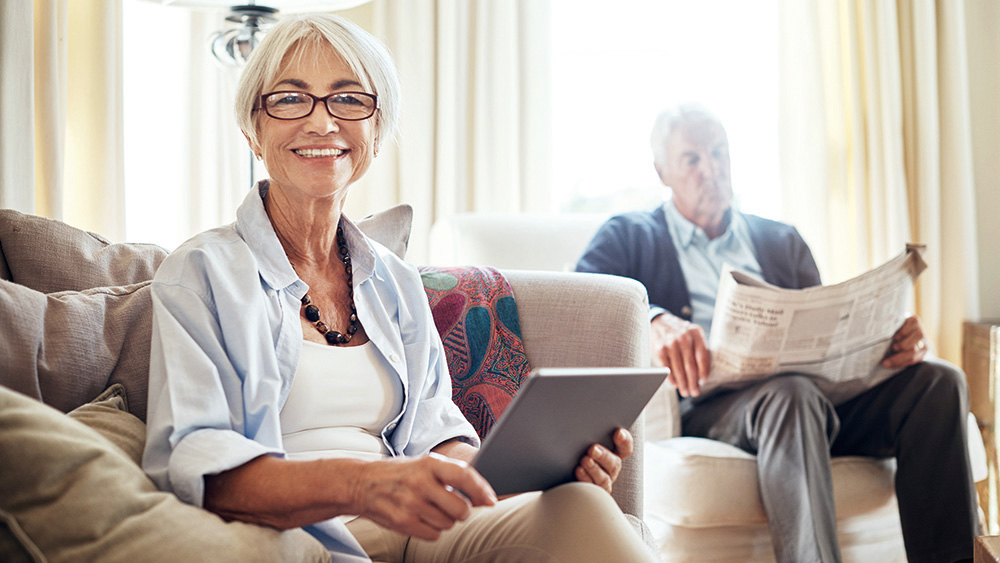 Portrait of a senior woman using a digital tablet while her husband reads a newspaper in the background