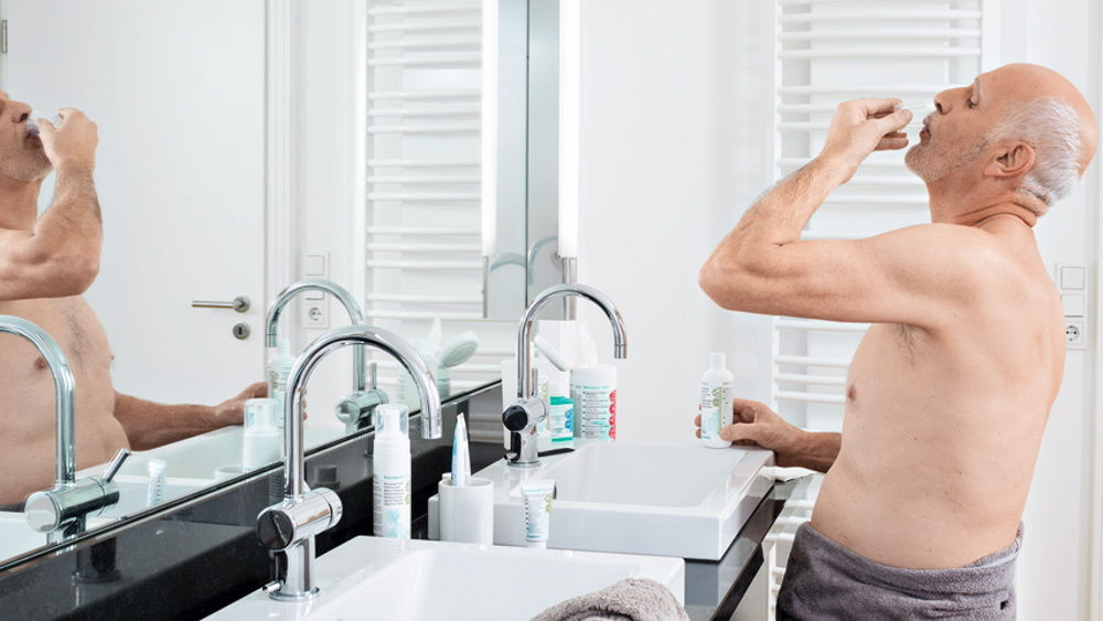 Älterer Mann spült zu Hause vor dem Badespiegel den Mund mit antimikrobiellen Mitteln 