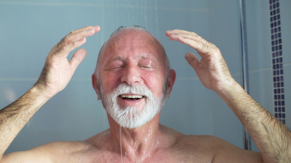 Ein älterer Mann steht in der Dusche und lächelt unter fließendem Wasser