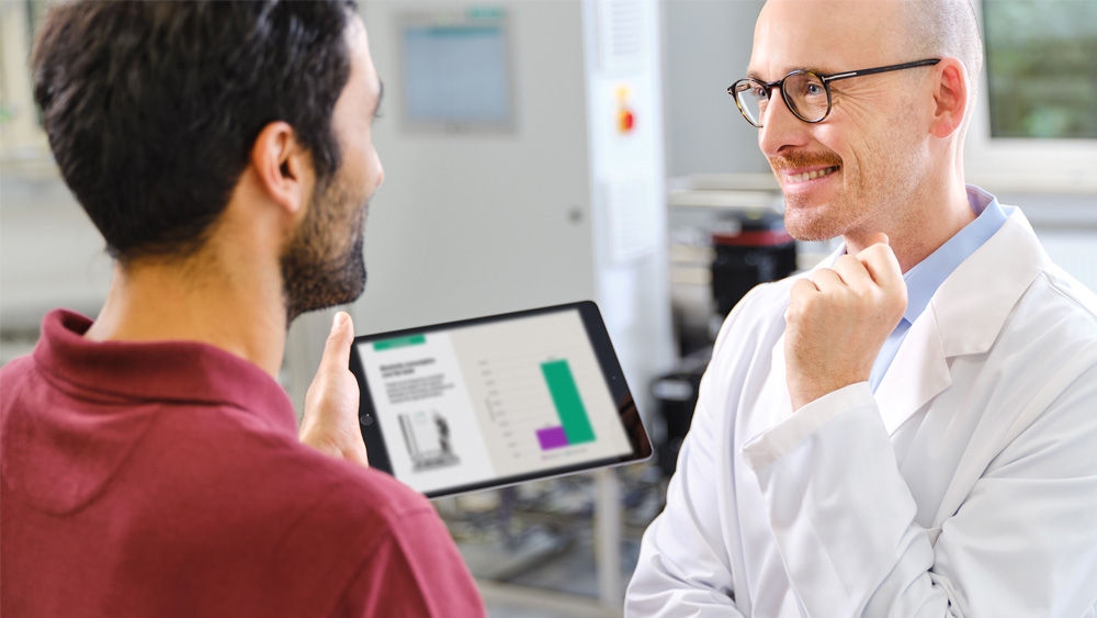 Zwei Personen, ein Nephrologe und ein Techniker, im Gespräch in einem Wasserbehandlungsraum. Der Nephrologe trägt einen weißen Laborkittel und der andere hält eine Tablette, die den Ressourcenverbrauch in der Dialyse anzeigt.