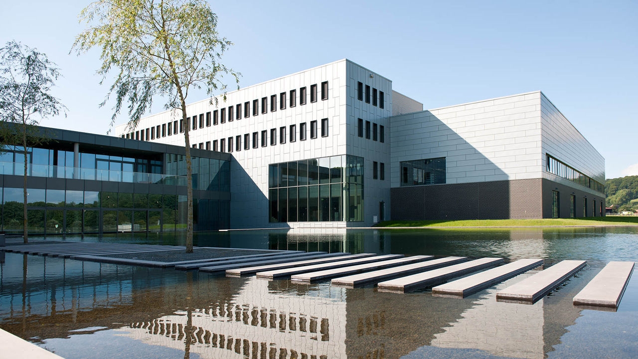 A modern glass building surrounded by a reflective pond with stepping stones, under a clear blue sky.