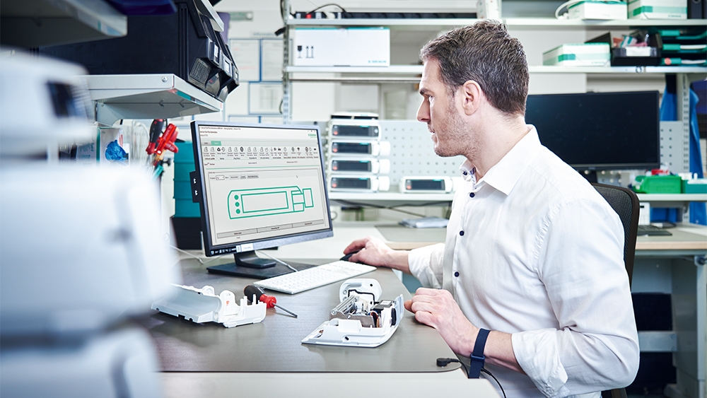Das Bild zeigt den technischen Kundendienst von B. Braun, der ein Infusionsgerät auf seinem Schreibtisch repariert, während er sich die Leistungsdiagnose auf seinem Computer ansieht.