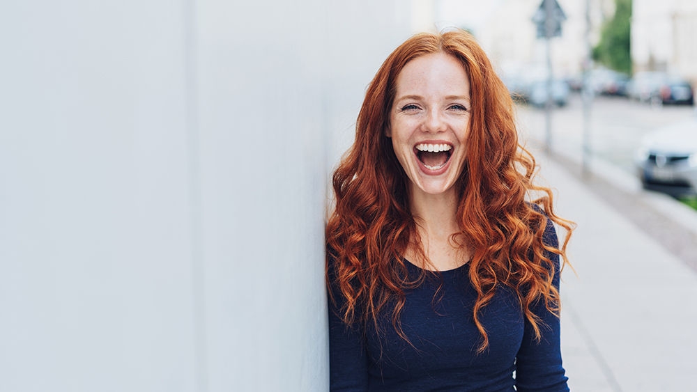 Cute young woman with a lovely sense of humour standing leaning against a white exterior wall with copy space in an urban street laughing at the camera