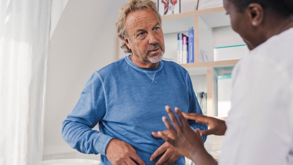 Ein mittelalter Mann stellt dem Arzt eine Frage, während er die rechte Seite seines Bauches hält, ein männlicher Arzt erklärt ihm etwas