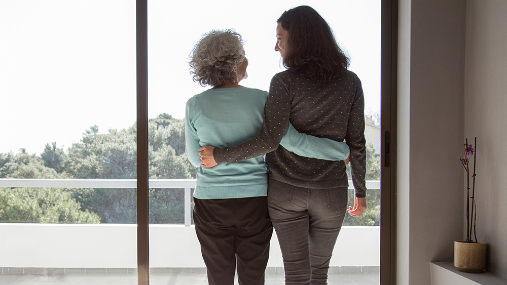 Rear view of happy mother and daughter standing embracing at window. Senior and mid adult women hugging and talking at home. Family relationships concept