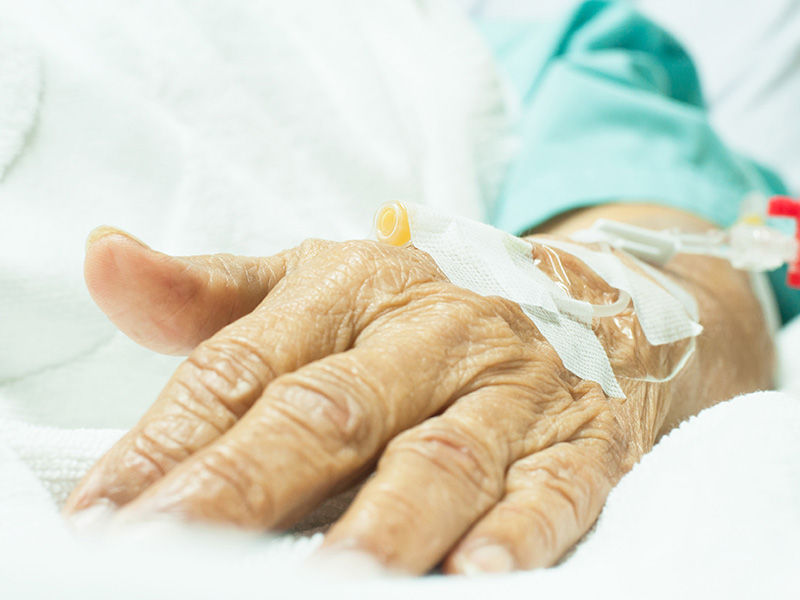 Geriatrische, faltige Hand liegt auf weißer Tagesdecke. Punktionsstelle am Handrücken. Angeschlossene Leitung mit weißer Schiebeklemme wird mit weißem Pflaster fixiert. Arm trägt grüne Krankenhauskleidung vom Ellenbogen nach unten.