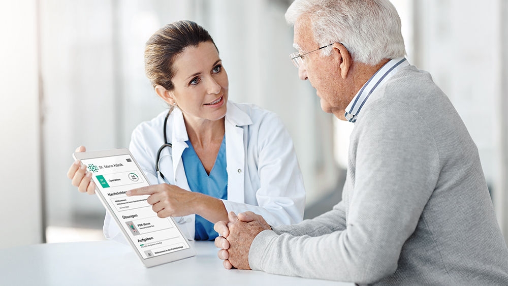 Ein Arzt sitzt an einem Tisch mit einem älteren Patienten und erklärt auf einem Tablet-Bildschirm Informationen über die ForPatientApp.