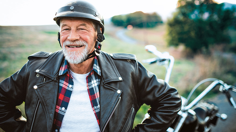 A cheerful senior man traveller with motorbike in countryside, standing., A cheerful senior man traveller with motorbike in countryside, s