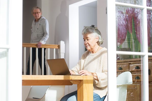 Eine ältere Dame sitzt am Fenster vor ihrem Laptop, im Hintergrund beobachtet sie ein älterer Herr.