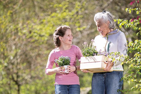 Eine ältere und eine jüngere Frau halten im Garten Blumensetzlinge in der Hand und lachen sich an.