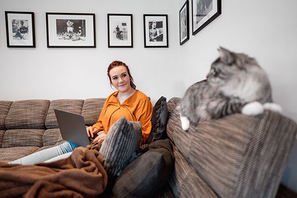 Eine junge Frau sitzt mit dem Laptop auf einer Couch und schaut ihre Katze an, die daneben liegt. 