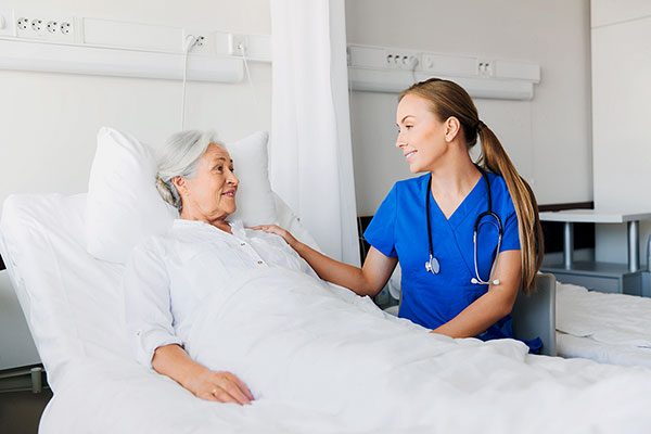 Eine ältere Frau liegt in einem Krankenhausbett während eine junge Krankenschwester neben ihr sitzt und sich mit ihr unterhält. 
