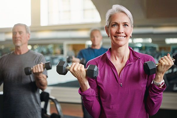 Eine ältere Frau mit Stoma ist in einem Fitnessstudio und hält zwei Gewichte in der Hand.