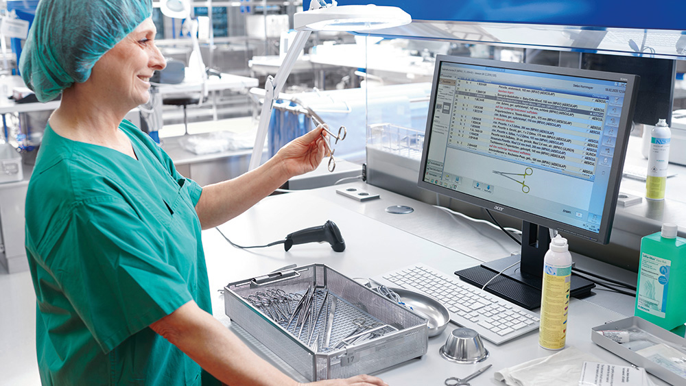 Medizinische Fachkraft in OP-Kleidung steht vor einem Computer, hält ein chirurgisches Instrument und vergleicht es mit einem Bild auf dem Bildschirm