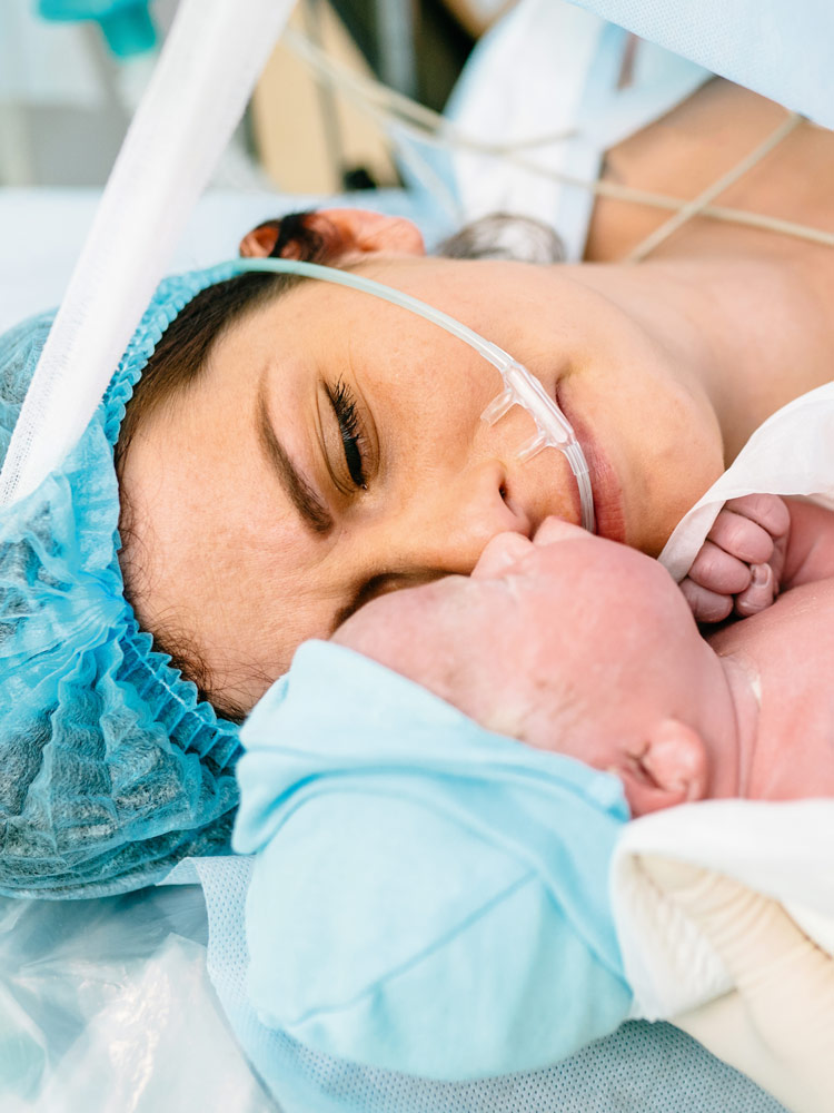 Eine Mutter liegt nach einem Kaiserschnitt im Operationssaal und betrachtet ihr neugeborenes Baby, das ihr eine Krankenschwester zeigt. Sie sieht erschöpft, aber auch erleichtert aus. Sie trägt eine OP-Mütze über ihren dunklen Haaren, das Neugeborene ist in ein weißes Tuch eingewickelt und trägt eine hellblaue Mütze.