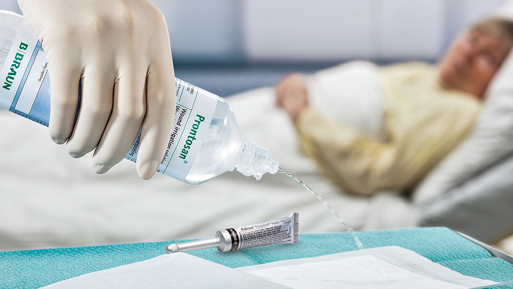 Eine Hand mit Handschuh hält eine Flasche Prontosan und drückt die Flüssigkeit auf ein großes Pflaster. Auf dem Tisch liegt eine Ashkina-Tube. Im Hintergrund liegt ein Patient im Bett.