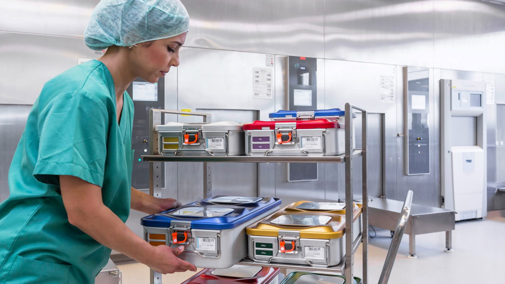 Eine weibliche medizinische Fachkraft in grüner OP-Kleidung steht in einem klinischen Umfeld und hält einen der gezeigten medizinschen Container.